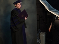 A male student wearing graduation attire and holding a diploma poses for a photo