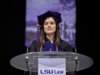 A female student wearing graduation attire speaks at a podium