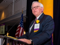 A man wearing a suit and tie talks at a podium with purple and gold banners in the background