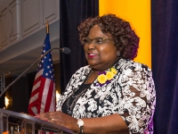 A woman talks at a podium with purple and gold banners in the background