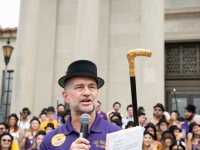 A man talks on a microphone with a group of people behind him on the steps of the LSU Law Center