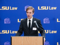 A male student wearing a suit and tie speaks at a podium with the LSU Law logo in the background