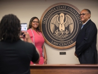 A female student wearing white cords and a man smile next to the LSU Law Center seal as a woman takes a photo on a phone