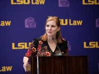 A woman wearing a black and red dress talks at a podium with the LSU Law logo in the background