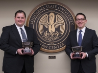 Two male students wearing suits and ties hold silver trophies next to the LSU Law Center seal