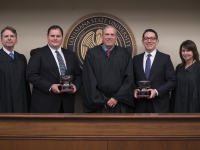Two male students wearing suits and ties hold silver trophies and pose with three judges next to the LSU Law Center seal