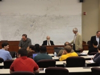 A group of male professors talk at the front of a classroom