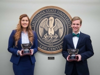A male and female student hold silver bowl trophies and smile for a photo next to the LSU Law Center seal